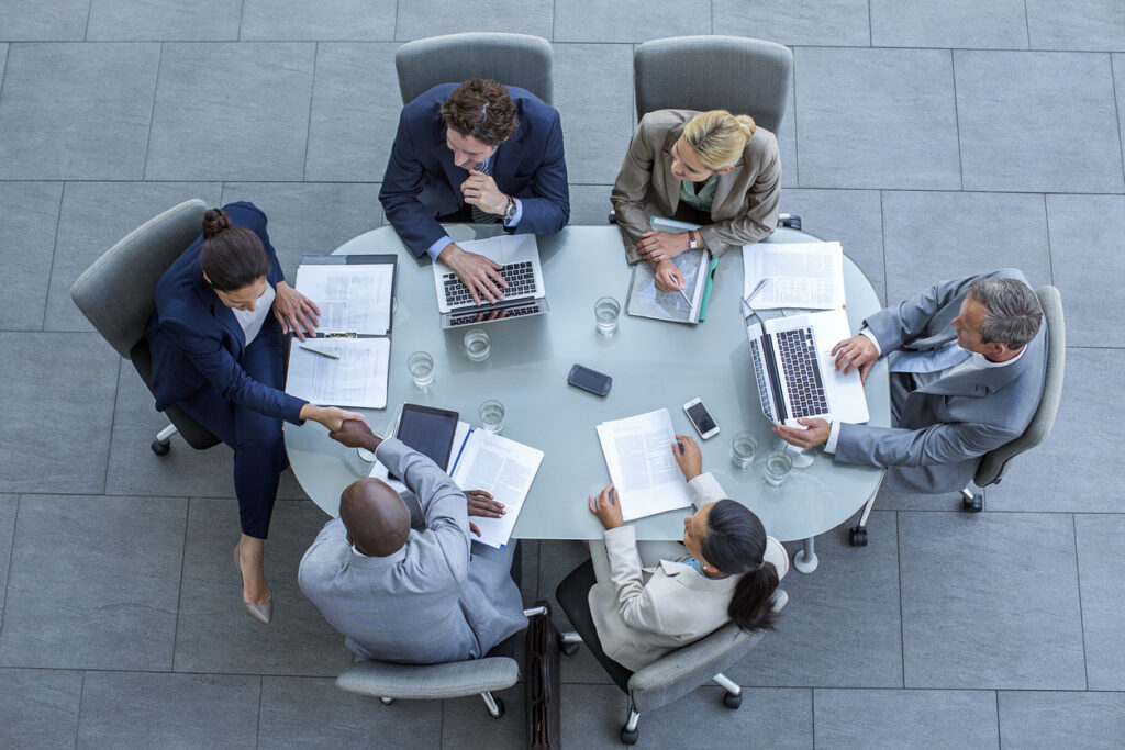 Six businesspeople sit around a round glass table with laptops, documents, and phones. Two are shaking hands, while others look at papers or screens—focusing on topics like cybersecurity and human resources strategies. The setting is a modern office with a tiled gray floor.