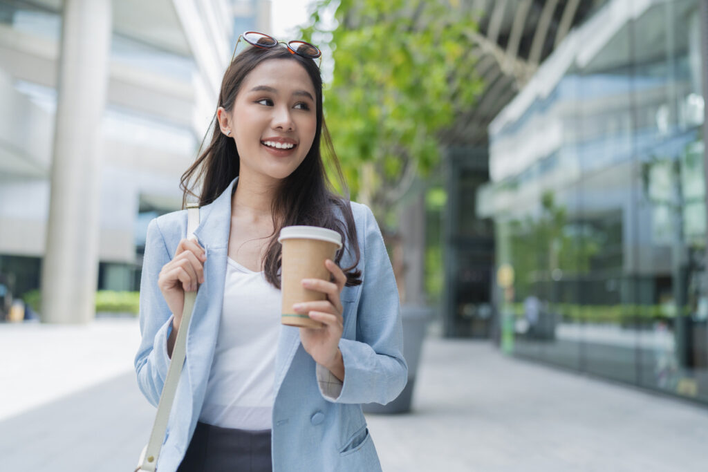 A young woman in a blue blazer holds a coffee cup and smiles while walking outdoors in a modern city area, with glass buildings and greenery in the background, embodying the confidence of a human resources professional navigating today’s dynamic business environment.