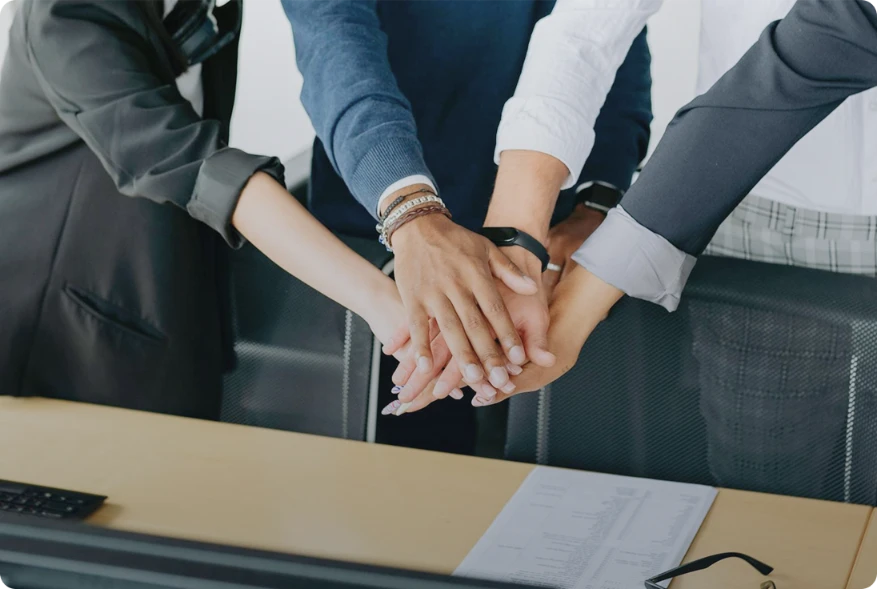 Five people standing around a desk stack their hands together in a gesture of teamwork and unity, demonstrating their shared commitment to cybersecurity. A document and keyboard are visible on the desk in front of them.