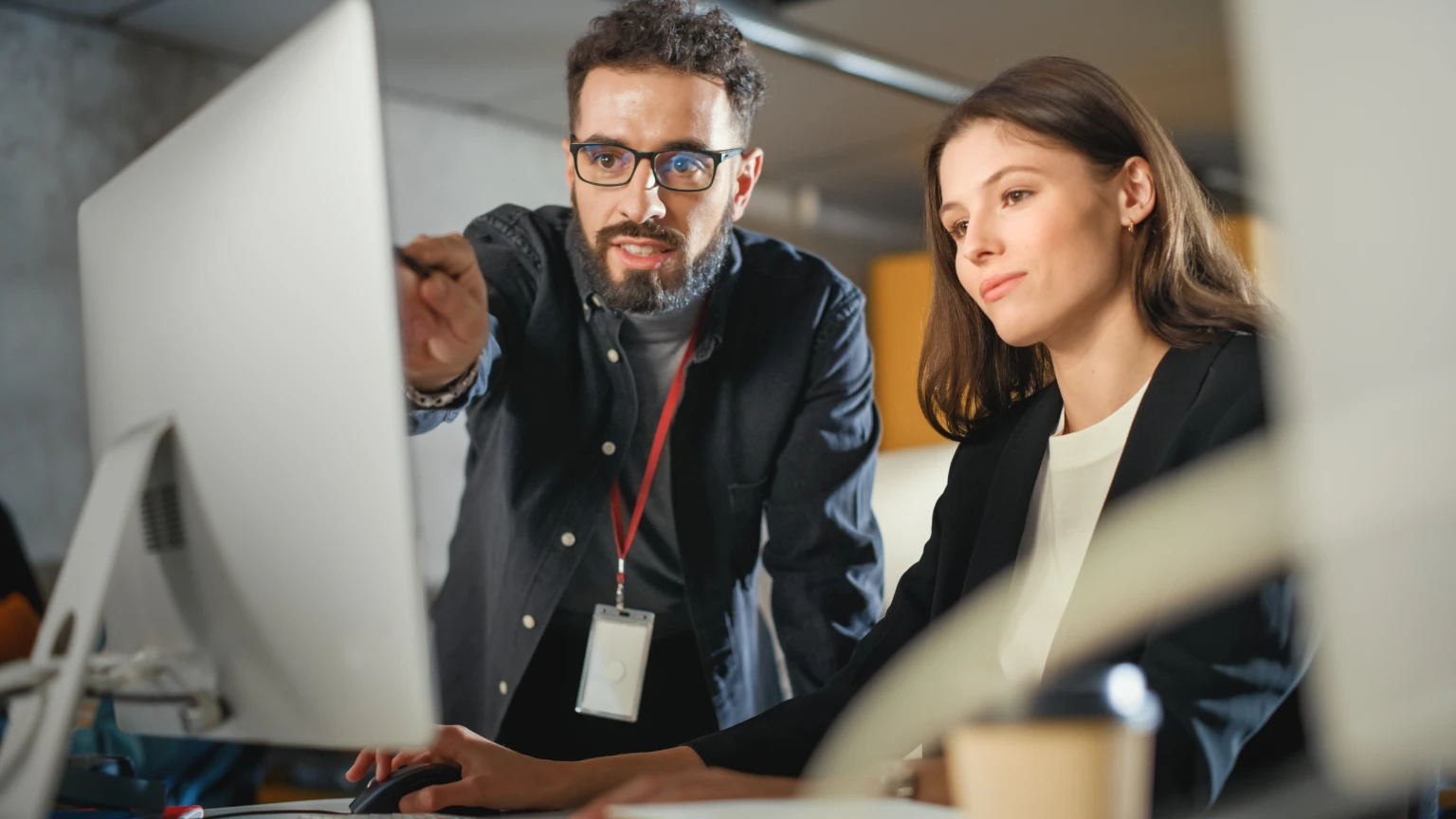 A man with glasses points at a computer screen while a woman sits next to him, both focusing on the monitor in a modern office setting as they discuss important cybersecurity measures.