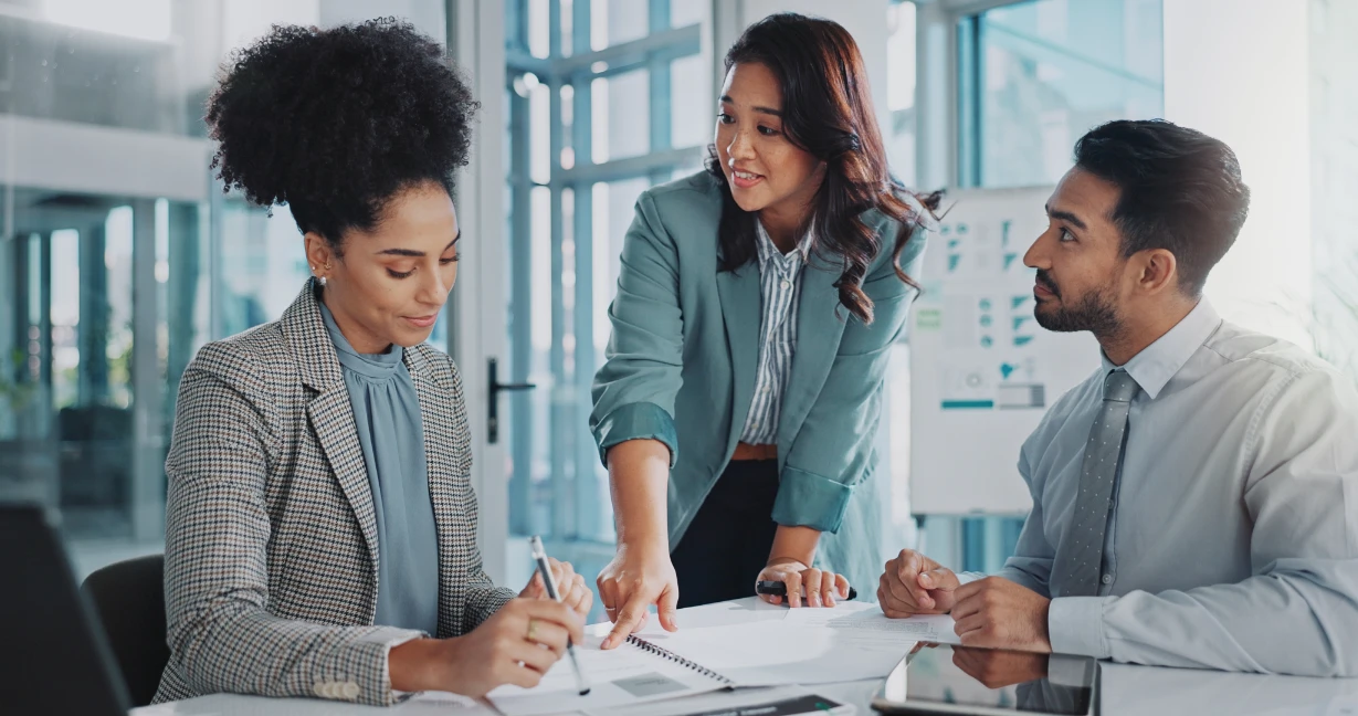 Three business professionals in an office discuss documents related to cybersecurity. One woman stands and points at paperwork, while a seated woman writes and a man listens attentively. Sunlight streams through large windows in the background.