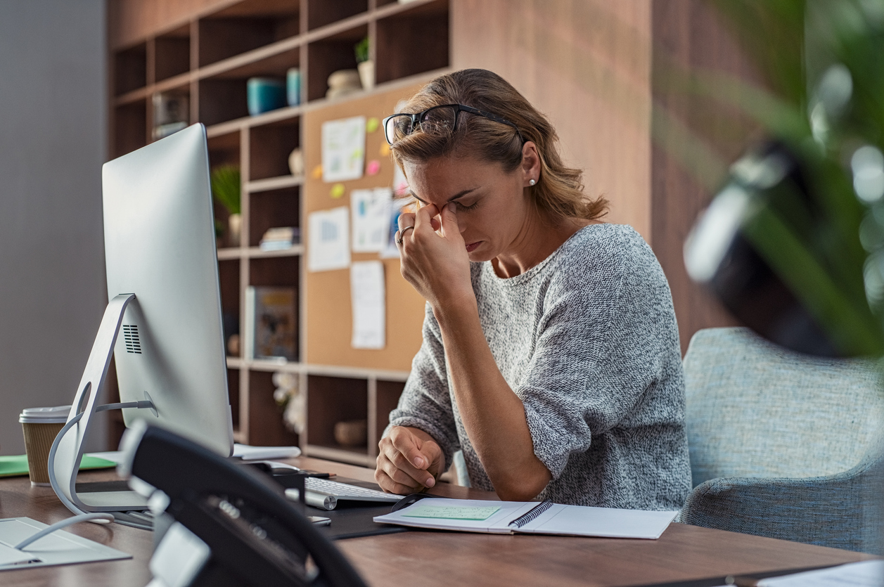 A woman sits at a desk in an office, holding her head and pinching the bridge of her nose, appearing stressed or fatigued amid paperwork, a computer, and office supplies—a scene reflecting the challenges often faced in human resources when managing business risk.
