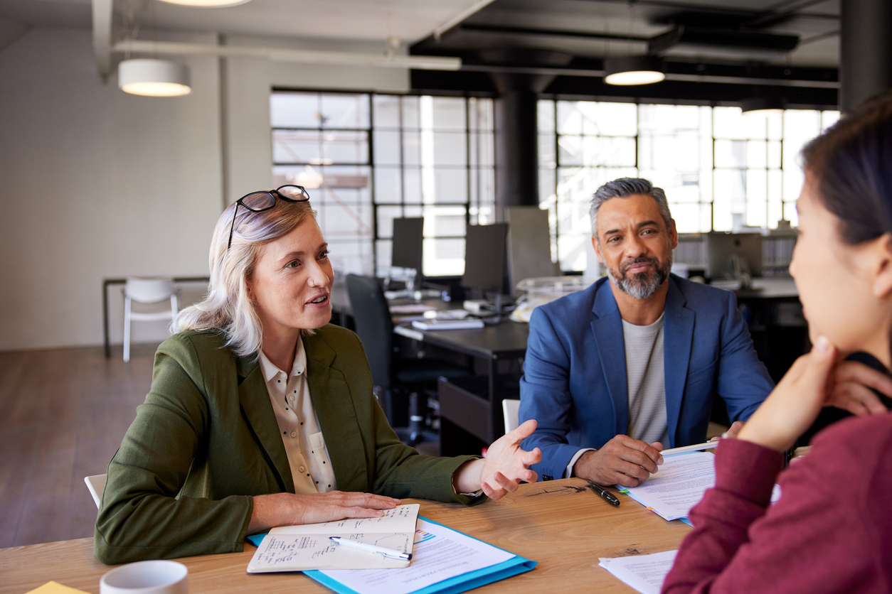 Two professionals, a woman and a man, sit across from another woman at a desk in a modern office, engaged in a discussion about business risk and human resources, with documents and notebooks on the table.