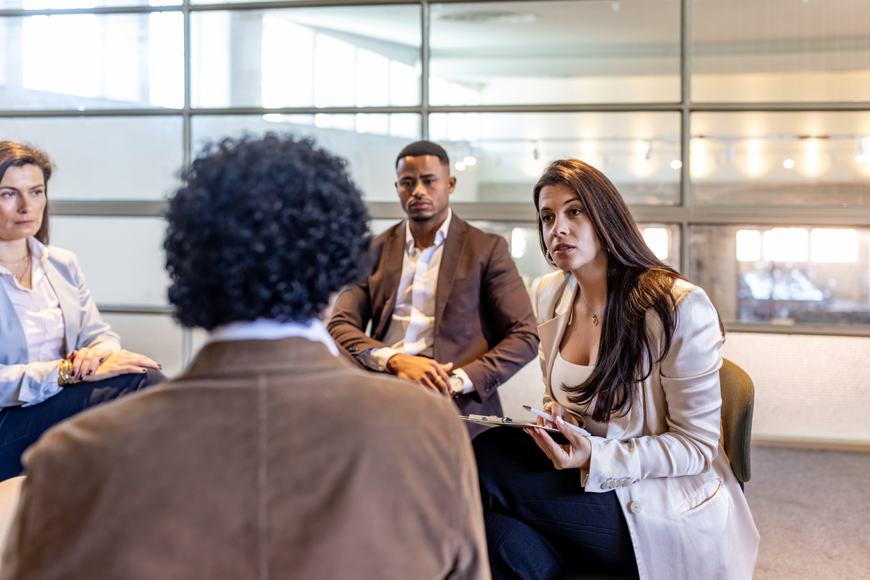 Four people in business attire sit in a modern office, engaged in discussion. One woman with long hair speaks, holding a notebook, while others listen attentively—perhaps strategizing on human resources initiatives or business risk management. Large windows are in the background.