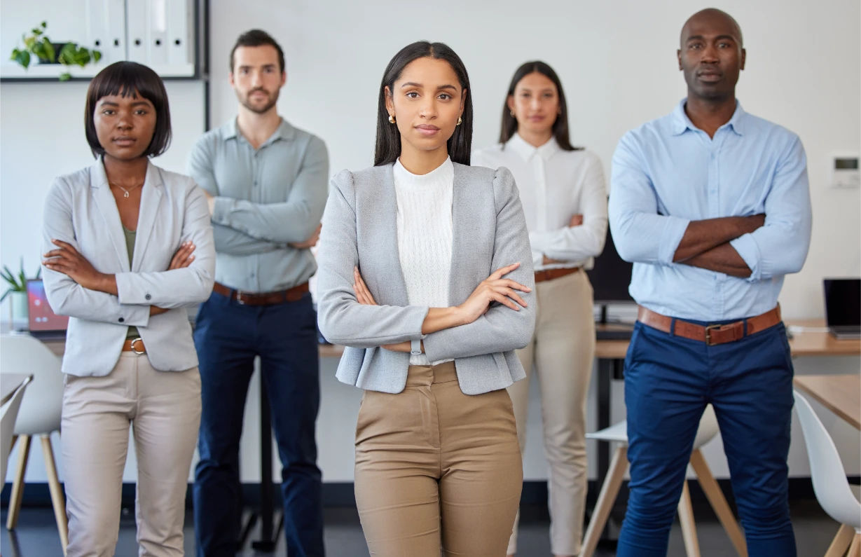 Five professionals stand confidently with arms crossed in an office setting, facing the camera. The group is diverse in gender and ethnicity, dressed in business-casual attire, suggesting teamwork and leadership.