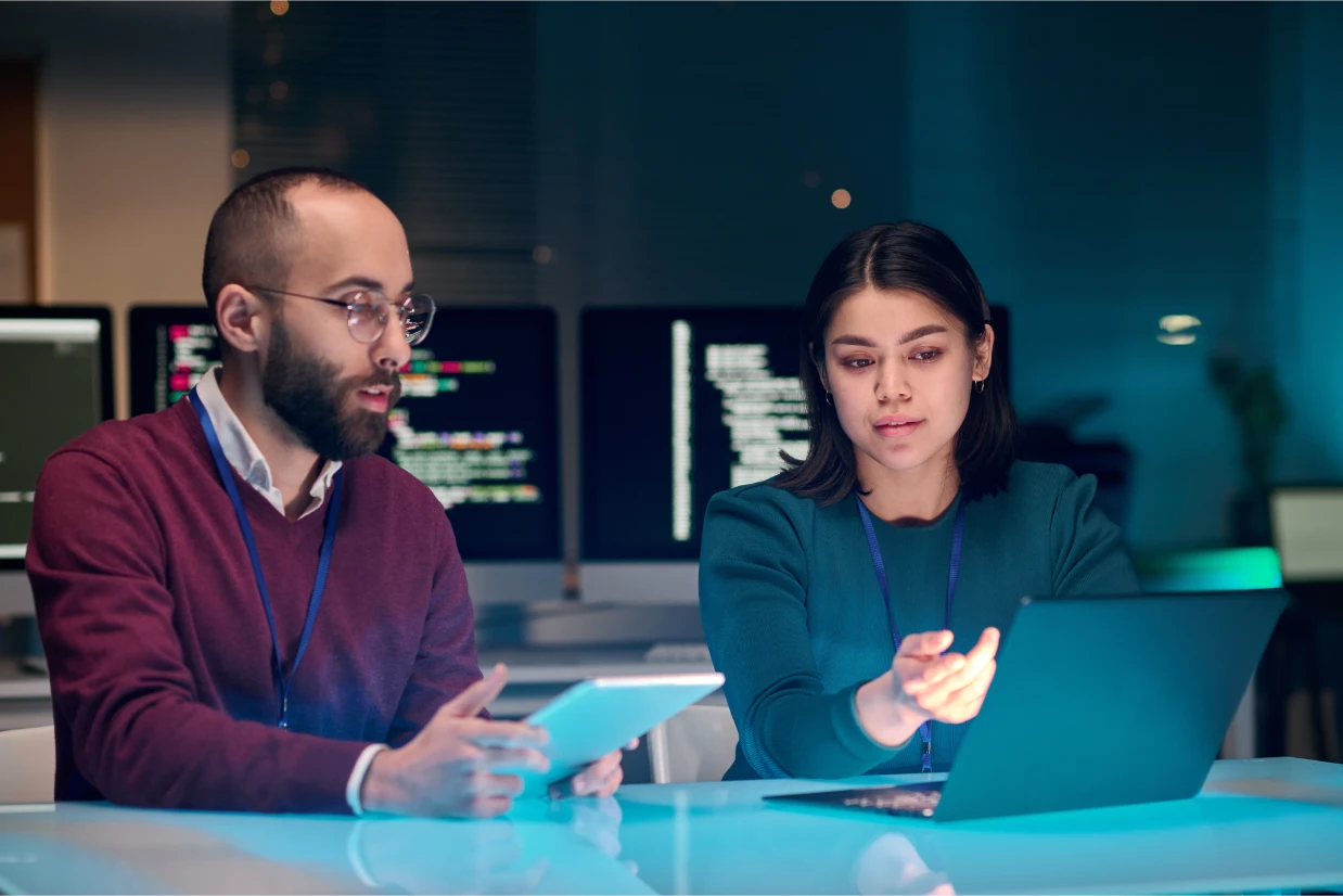 Two people sit at a desk in an office at night, working on a laptop and a tablet. Computer monitors with code are in the background, and both appear focused on their work.