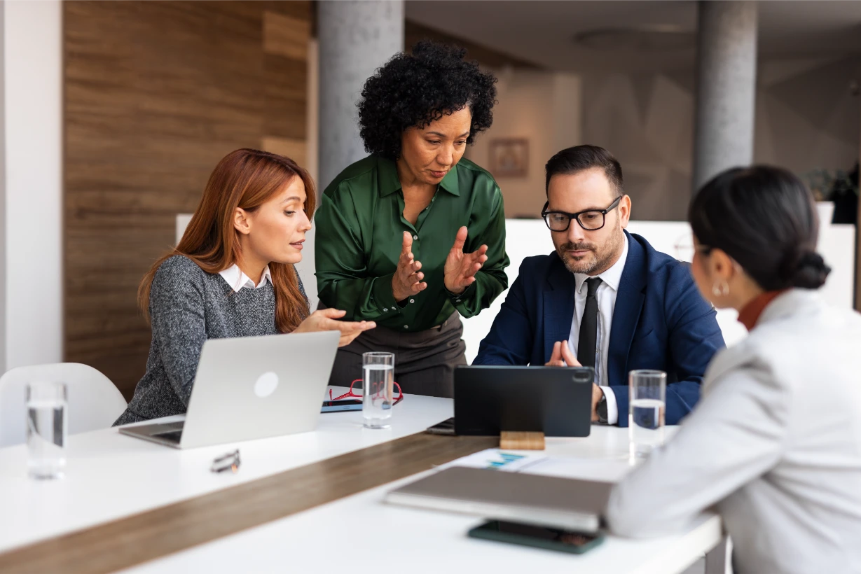Four professionals in business attire sit at a table with laptops, papers, and water glasses, engaged in a discussion. One woman stands, gesturing with her hands, while the others listen and look at a tablet.