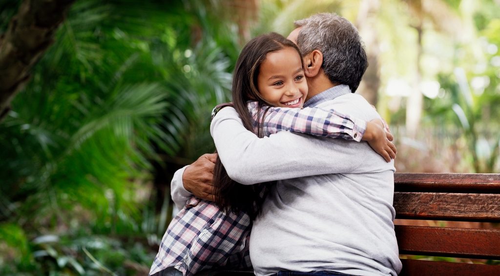 A young girl hugs an older man on a bench outdoors, both smiling, surrounded by green foliage—a scene that highlights the importance of staying alert to child grooming warning signs, even in seemingly innocent moments.