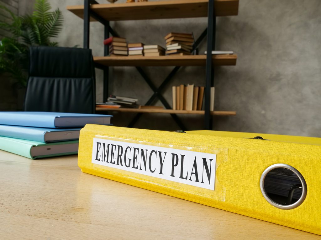 A yellow binder labeled Emergency Plan sits on a desk, ready to guide actions during the critical first 24 hours of a cyber incident. Behind it are stacked binders, an empty chair, and a bookshelf with books and plants, creating an office setting.
