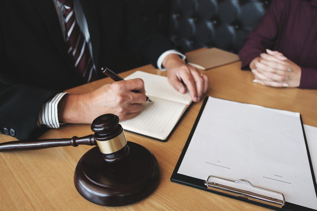 A person in a suit writes in a notebook at a desk with a gavel and a clipboard, possibly documenting employment practices liability claims, while another person sits across from them with hands folded.