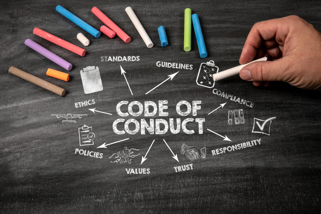 A hand holds chalk near a chalkboard with Code of Conduct in the center, surrounded by words like ethics, trust, policies, values, guidelines, standards, compliance, and responsibility. Notably, terms such as signs of grooming behavior appear among these principles. Colored chalk lies nearby.