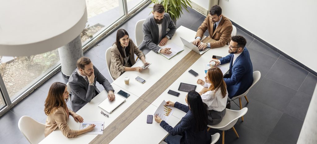 Eight people in business attire sit around a white conference table in a modern office, discussing documents and using laptops and tablets as they collaborate on developing a cyber incident response plan during their meeting.
