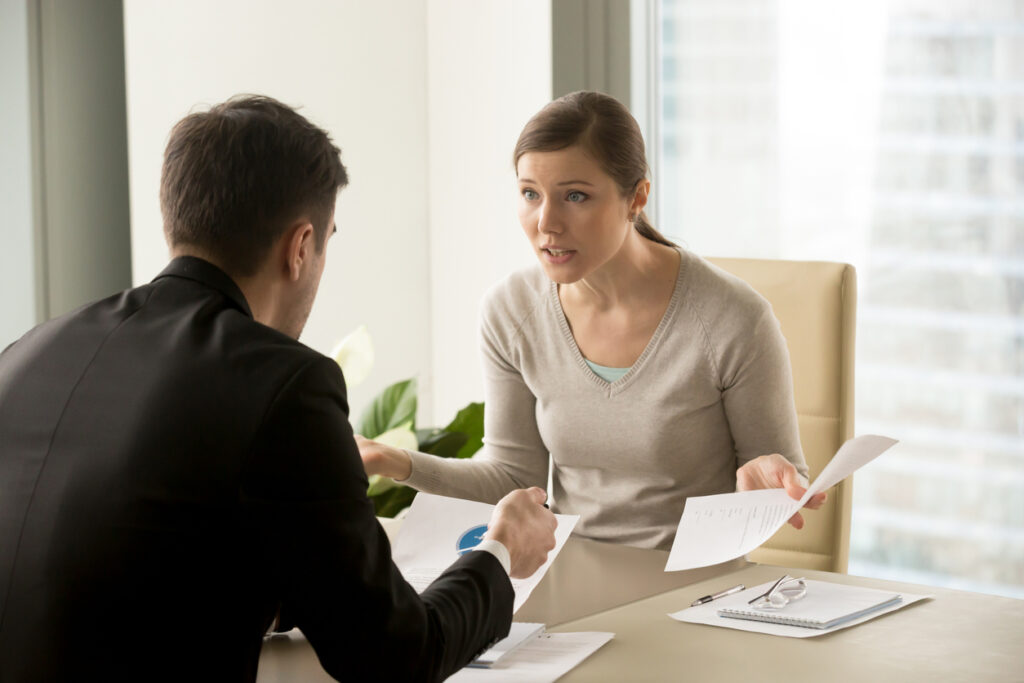 Two people sit at a desk in an office, engaged in a serious conversation that hints at workplace investigations. The woman holds a document, gesturing emotionally, while the man listens intently, partially turned away from the camera.