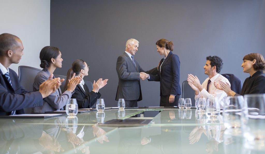 A new board member and a colleague shake hands at the head of a conference table while others applaud.